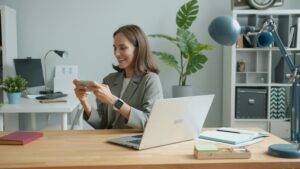Start 12 Prokrastination einer Frau am Arbeitsplaz. Sie guckt aufs Handy und sitzt vor ihrem Laptop.